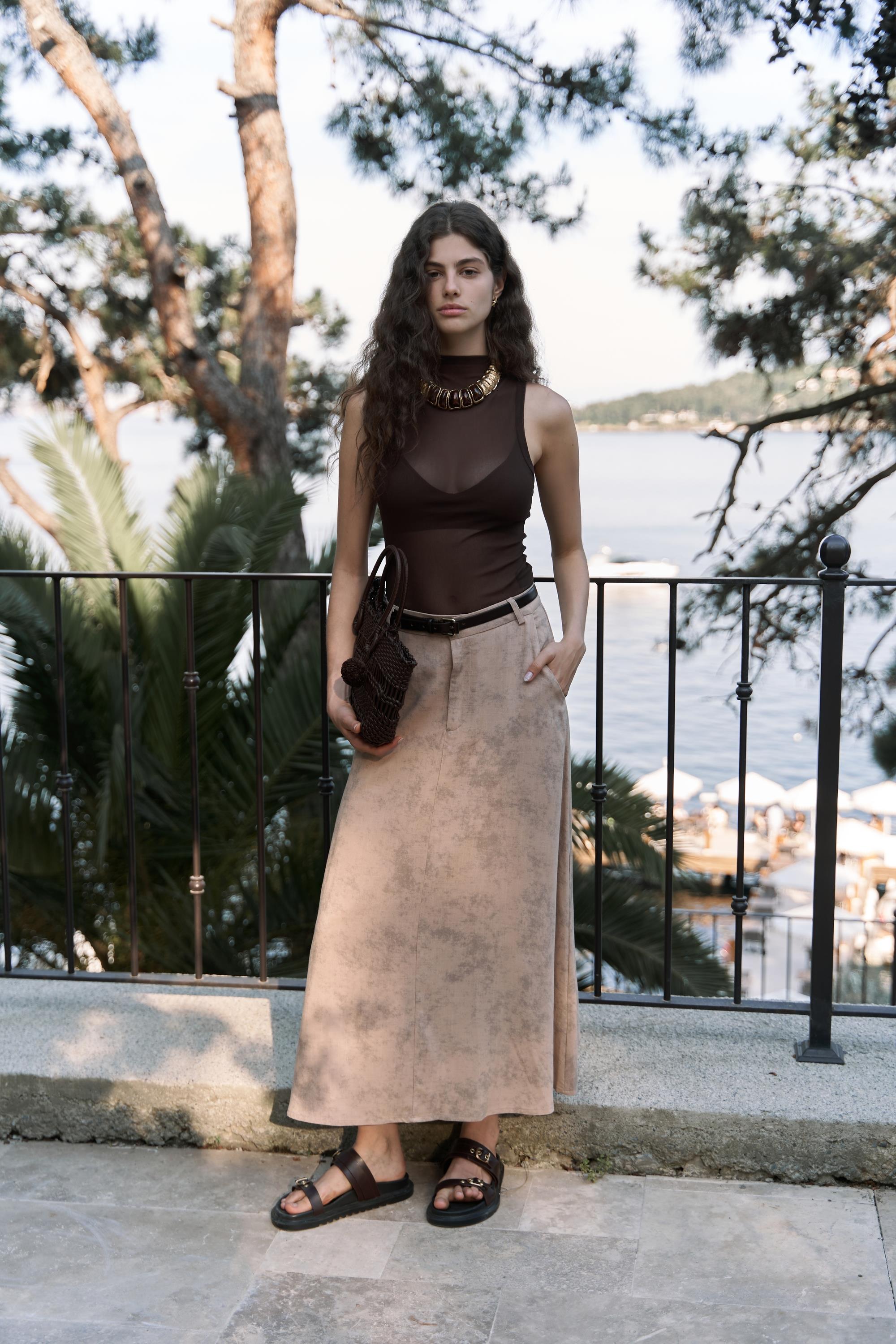Woman wearing a sheer top and beige batik printed skirt outdoors with a sea in the background