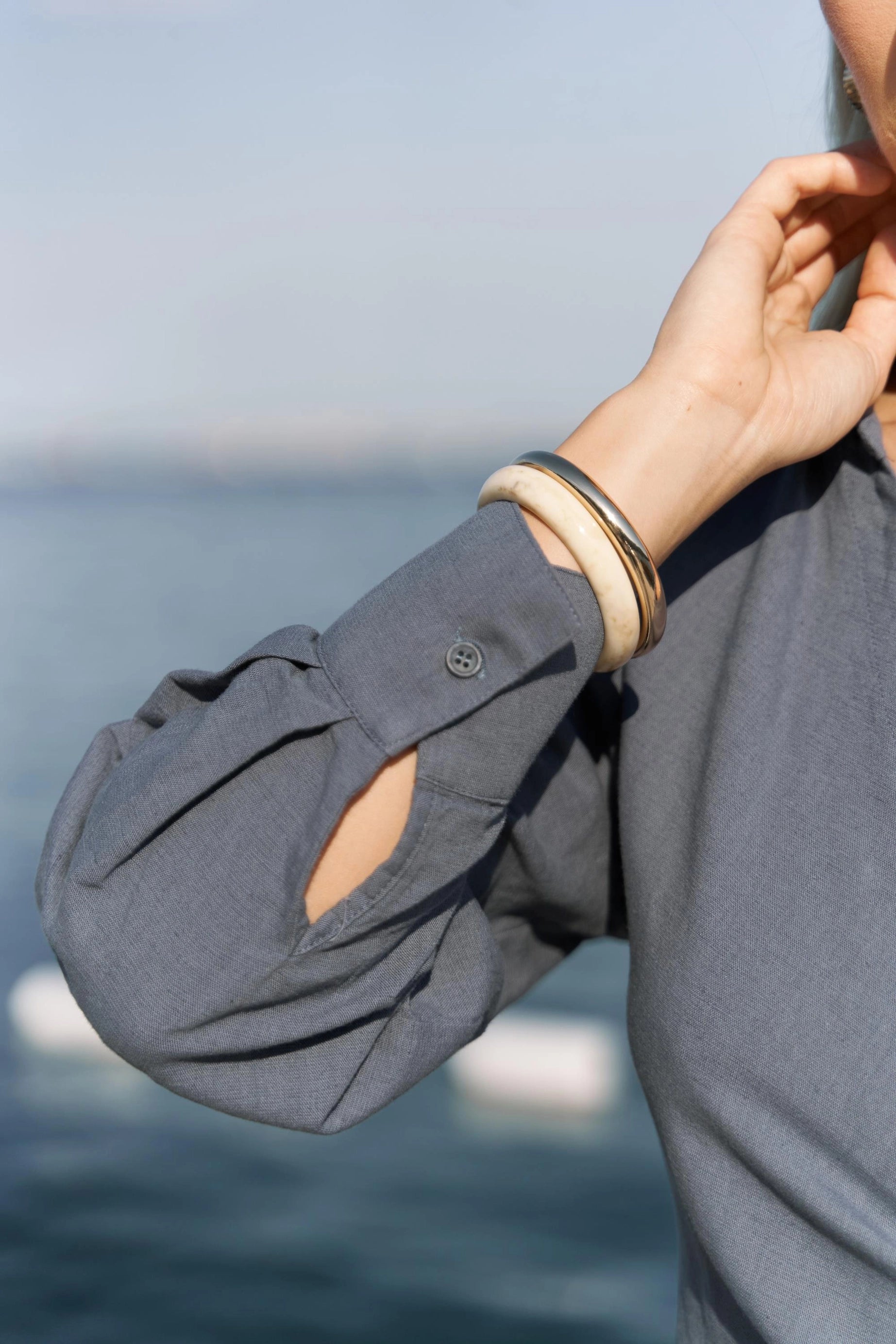 A woman wearing a indigo cotton dress with rolled-up sleeve, hand adjusting it, against a blurred ocean background.