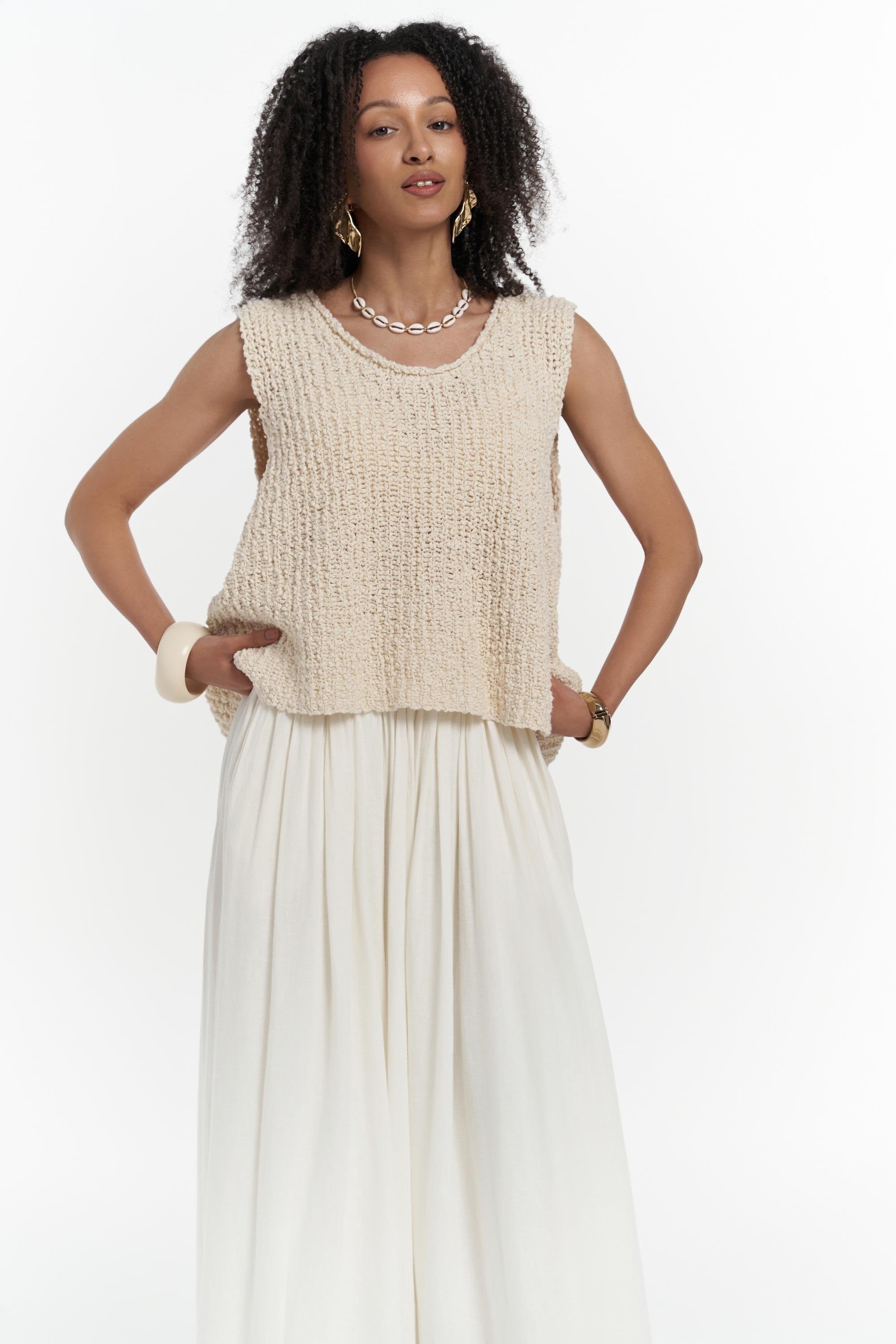 Woman wearing a beige knitted sleeveless top and white pants on a white background
