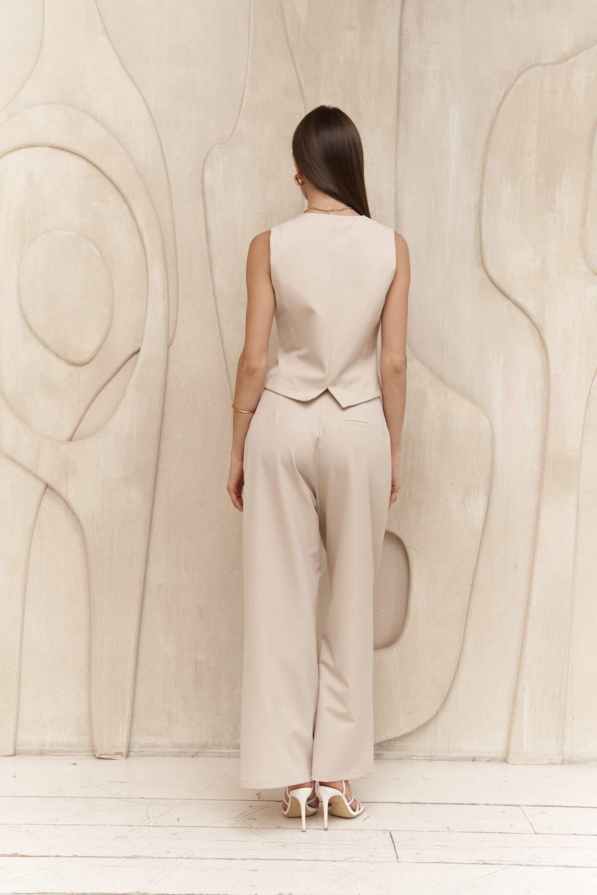Woman wearing a beige sleeveless top and wide-leg pants standing against a textured beige wall.