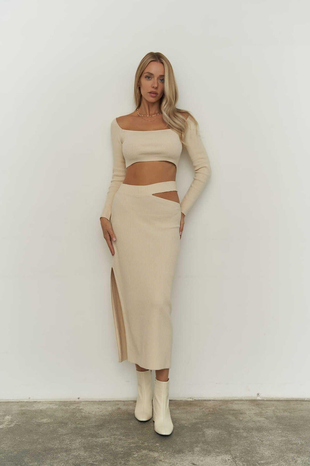 Woman wearing a beige ribbed knit outfit with a slit skirt against a white wall.