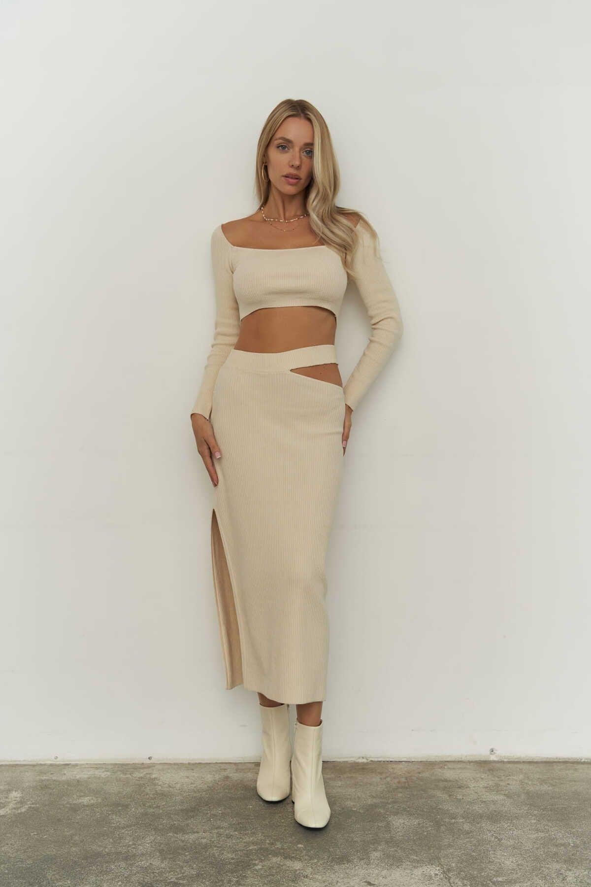 Woman wearing a beige ribbed knit outfit with a slit skirt against a white wall.